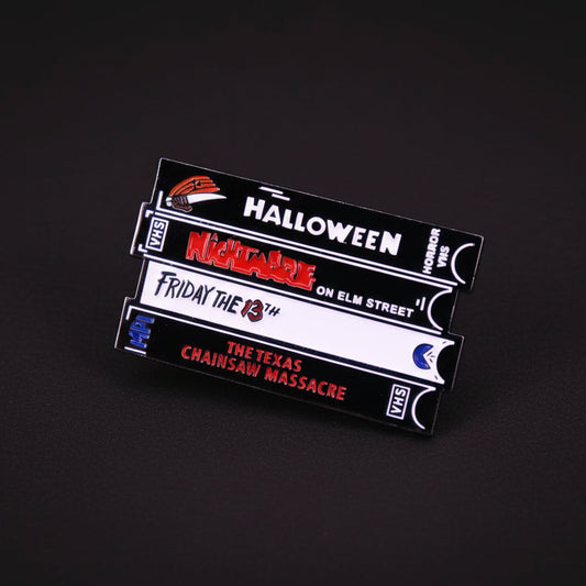 Enamel pin of a set of horror-themed VHS tapes on a dark background. The tape titles include Halloween, Nightmare on Elm Street, Friday the 13th, and The Texas Chainsaw Massacre