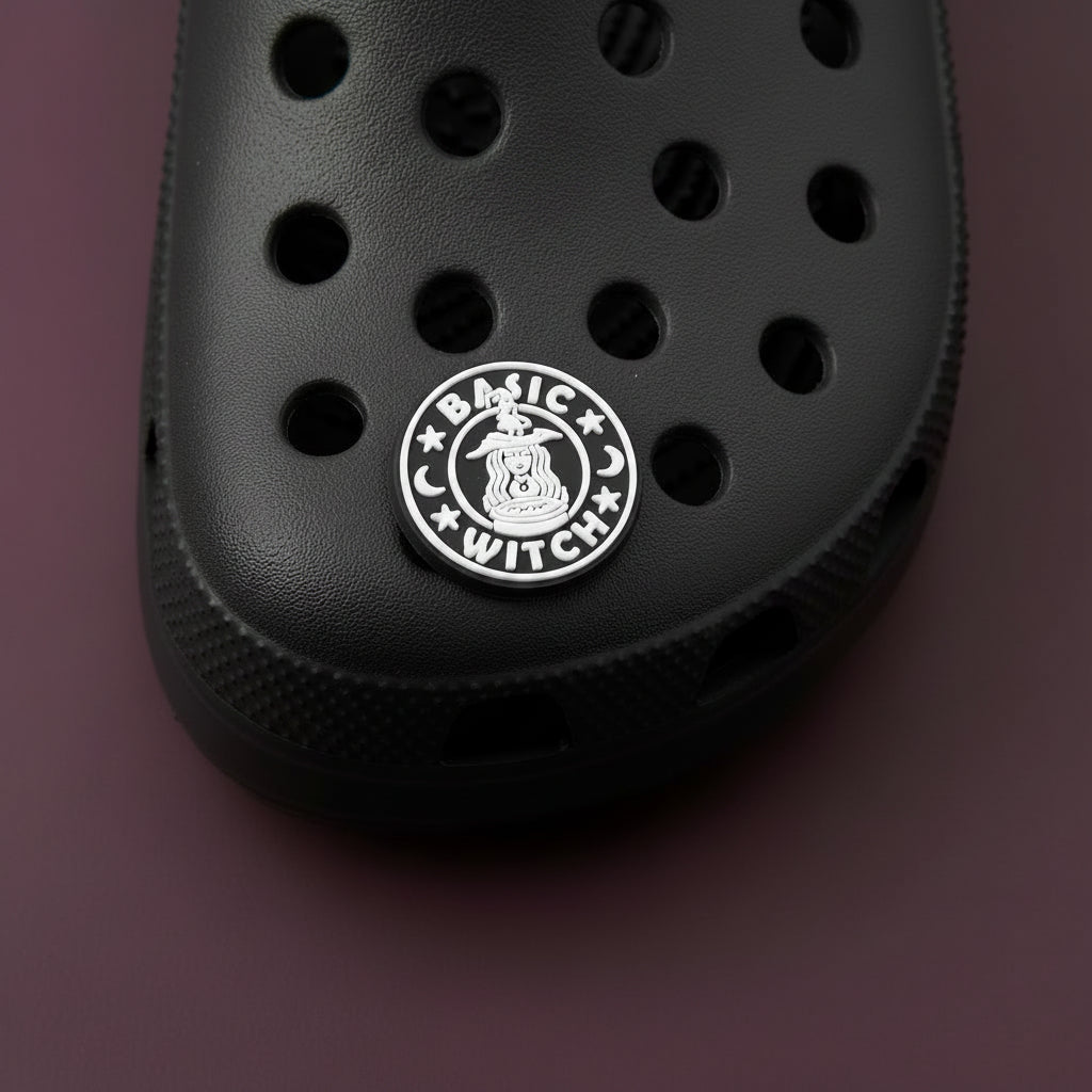 Black circular clog charm with white imaging and text that resembles the Starbucks mermaid logo, but instead of a mermaid, it is a witch above a cauldron with a pointy witch hat. The image is surrounded by the phrase "Basic Witch" and two crescent moons and stars.