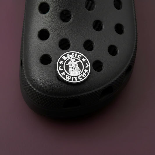 Black circular clog charm with white imaging and text that resembles the Starbucks mermaid logo, but instead of a mermaid, it is a witch above a cauldron with a pointy witch hat. The image is surrounded by the phrase "Basic Witch" and two crescent moons and stars.